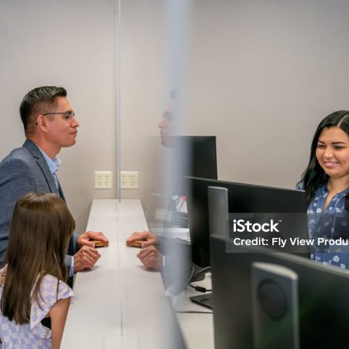 A cheerful female customer service representative assists a Latin American man and his elementary age daughter at the reception desk of a medical office or bank.